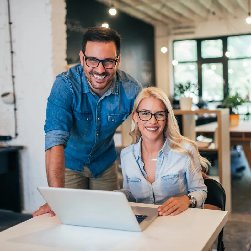 Two smiling professionals in a modern office collaborate at a laptop, showcasing teamwork and innovation.