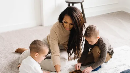 Woman and two young boys playing a board game together while sitting on a carpet in a bright living room.