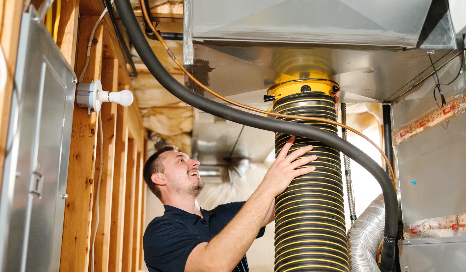 Technician inspecting flexible ductwork in unfinished basement HVAC system with exposed wooden framing and insulation.
