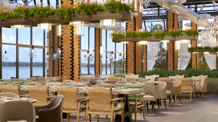Modern restaurant interior with chandeliers, greenery, and large windows overlooking the waterfront.