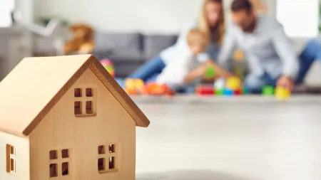 Wooden toy house in foreground with blurred family playing together in bright living room background